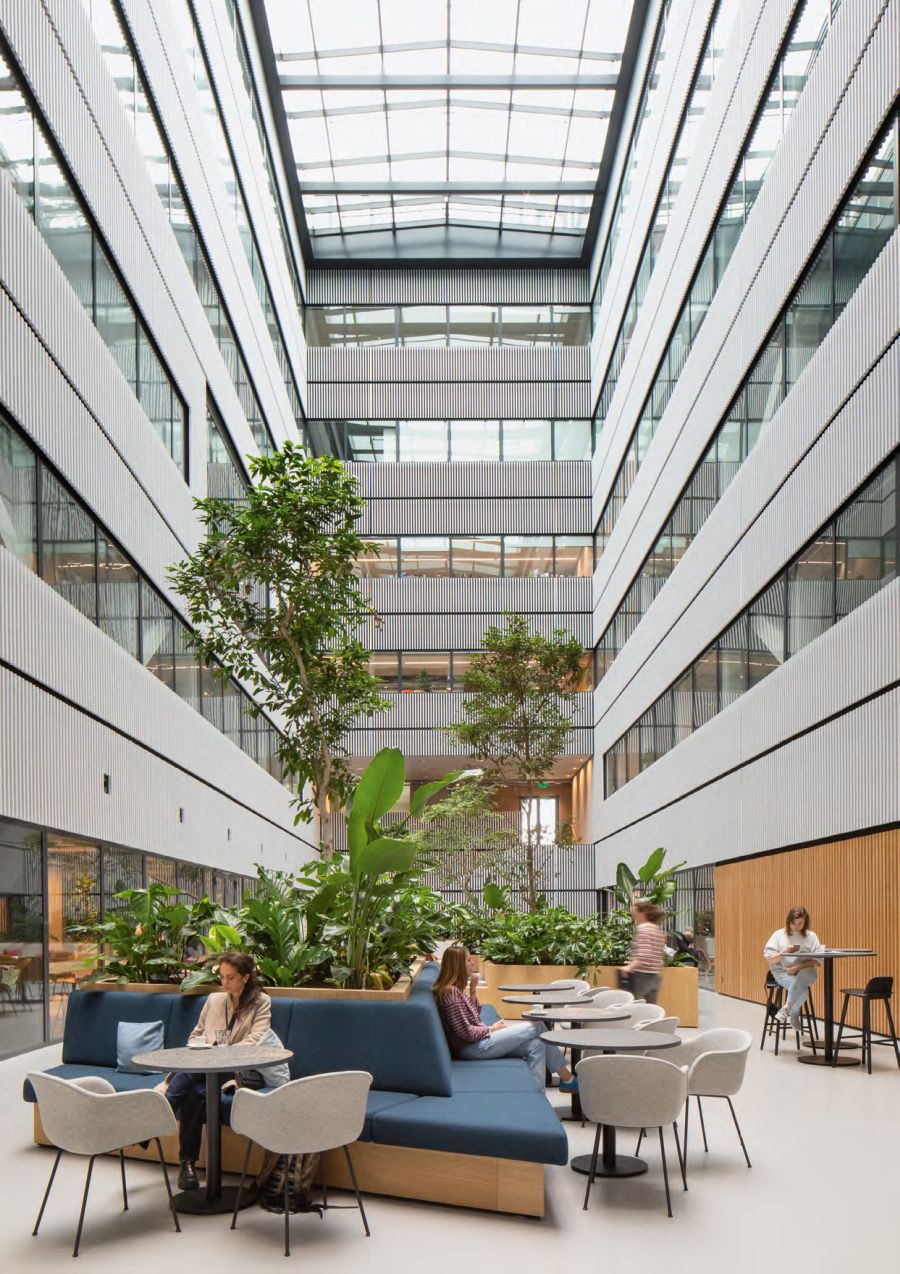 Das Bild zeigt das weitläufige, helle Atrium eines modernen Bürogebäudes. Das Atrium erstreckt sich über mehrere Stockwerke und wird von einem Glasdach beleuchtet. Entlang der oberen Stockwerke befinden sich Bürogänge mit weiß verkleideten Balkongeländern und Glasfenstern. Im Erdgeschoss ist ein gemütlicher, begrünter Aufenthaltsbereich eingerichtet. Große Topfpflanzen und Bäume säumen den Bereich. Mehrere Sitzgruppen sind vorhanden, darunter eine große, blaue, geschwungene Couch mit kleinen runden Tischen sowie separate Bistrotische mit Stühlen. Auf der linken Seite sitzt eine Frau auf der blauen Couch. In der Mitte des Bildes sitzt eine weitere Person auf der Couch. Im Hintergrund rechts sitzt eine Frau auf einem Barhocker an einem hohen Tisch und liest. Die Atmosphäre wirkt entspannt und einladend, wie eine moderne Kaffeebar oder Loungebereich in einem Bürokomplex.