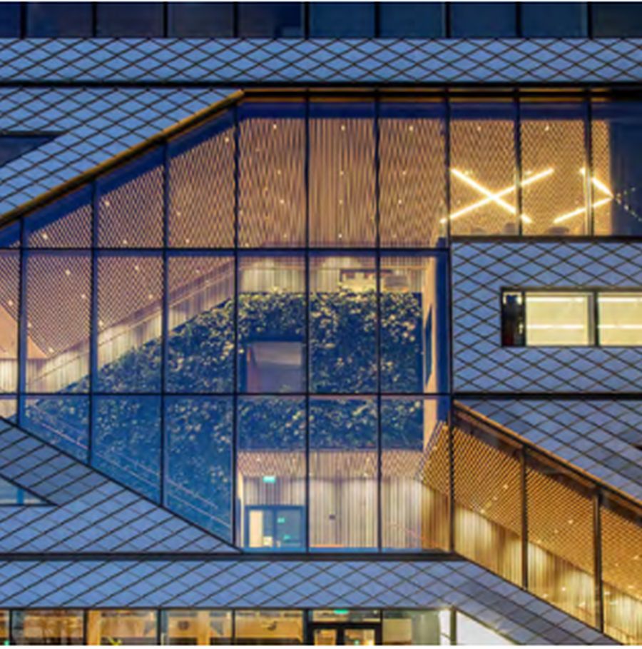 Diese Ansicht zeigt einen Teilausschnitt der Fassade des Gebäudes Matrix ONE bei Dämmerung. Die Fassade kombiniert helle, rautenförmige Aluminiumplatten mit einer markanten Glasfront. Große, diagonal verlaufende Glasflächen bilden ein geometrisches Muster, das den Verlauf der inneren Haupttreppe nach außen spiegelt. Das Gebäude strahlt von innen ein warmes, gelbliches Licht aus. Durch die Glasfassade ist der hell erleuchteten Innenbereich sichtbar. Schrägen und Handläufe zeigen, dass in diesem Bereich im Innenraum Treppen verlaufen. Hinter dem Glas werden eine hohe grüne Pflanzenwand und lamellenartige Wandverkleidungen sichtbar. Im oberen rechten Fensterbereich leuchtet eine markante X-förmige Lichtstruktur. Die Architektur wirkt durch den Kontrast von technischem Metall, transparentem Glas und organischem Grün hochmodern. Der untere Bildbereich zeigt den beleuchteten Eingang und weitere Innenräume. Die gesamte Komposition betont die offene, nachhaltige Bauweise des Forschungszentrums.