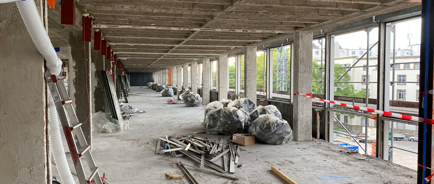 Foto aus dem Inneren eines unfertigen Gebäudes auf einer Baustelle: Der Raum besteht aus Betonwänden und -decken mit sichtbaren Stahlträgern, mehrere Fenster lassen Tageslicht herein. Auf dem Boden liegen Bauabfälle wie Metallstangen, Plastikfolien und weitere Materialien sowie ein Stapel schwarzer Müllsäcke – Anzeichen von selektivem Rückbau und Materialtrennung. An einigen Stellen sind rote und weiße Warnbänder gespannt. Die Perspektive führt von einem Raumende in die Tiefe des Korridors. Kein Text oder Bildnachweis sichtbar.