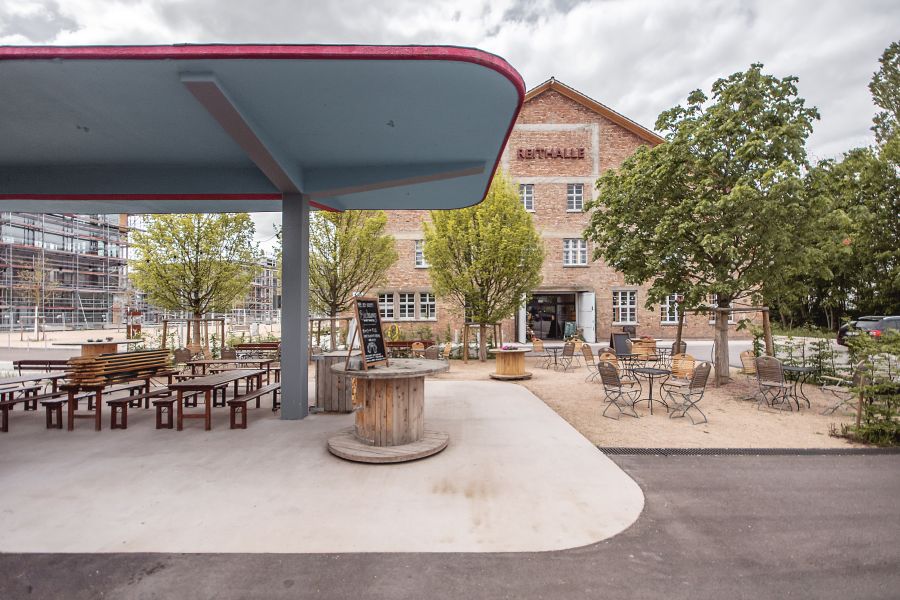 Zu sehen ist der Außenbereich vor dem Backsteingebäude mit der Aufschrift „REITHALLE“. Im Vordergrund links schützt ein großes, flaches, hellblaues Vordach mit roter Kante, vermutlich das Dach einer früheren Tankstelle, eine Terrasse mit Holztischen und Bänken. Mittig steht ein Stehtisch aus einer hölzernen Kabeltrommel mit einer Tafel (vermutlich Speisekarte). Rechts davon erstreckt sich ein Kiesplatz mit filigranen Metallstühlen und Tischen unter jungen Bäumen. Die Reithalle, ein historisches Gebäude, ist im Hintergrund zu sehen und hat helle Sprossenfenster sowie einen großen Eingang. Die Atmosphäre wirkt modern-rustikal und einladend.