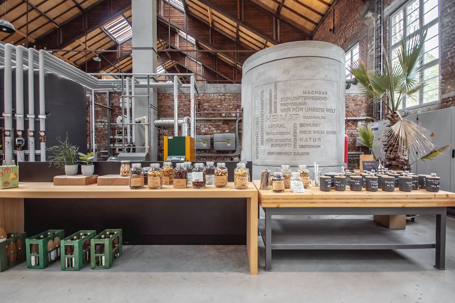 Das Bild zeigt eine modern gestaltete Verkaufs- oder Ausstellungsfläche im Innenraum der Reithalle. Der Raum hat hohe Decken und einen sichtbaren Dachstuhl. Im Vordergrund stehen zwei lange Holztische nebeneinander. Darauf befinden sich zahlreiche Glasgefäße und dunkle Dosen, gefüllt mit Trockenwaren oder Gewürzen. Unter dem linken Tisch stehen grüne Getränkekisten. Im Hintergrund dominiert ein großer, grauer Betonzylinder, der mit verschiedenen Schlagworten wie „HEIMAT“, „NATUR“ und „ZUKUNFT“ beschriftet ist. Links davon sind industrielle Rohrleitungen und Anlagen vor einer Backsteinwand zu sehen. Rechts bringt eine große Palme einen grünen Akzent in das industrielle Ambiente. Ein großes Sprossenfenster lässt Tageslicht herein. Die Atmosphäre wirkt durch die Kombination aus Holz, Sichtbeton und Ziegelsteinen modern und nachhaltig.