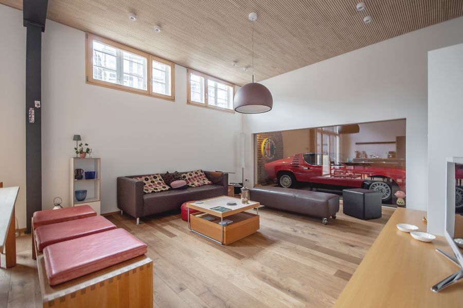 Das Bild zeigt ein weitläufiges, modernes Wohnzimmer: Hinter einer großen Glasfront ist ein klassischer roter Sportwagen in einer Art internen Garage geparkt, der als zentrales Designelement fungiert. Die Einrichtung ist minimalistisch und hochwertig: Ein dunkles Ledersofa mit gemusterten Kissen steht an der weißen Wand. Davor befindet sich ein quadratischer Couchtisch in Holzoptik. Verschiedene Sitzgelegenheiten wie massive Holzbänke mit roten, quadratischen, flachen Kissenund ein dunkler Ottoman ergänzen den Raum. Helles Eichenparkett und eine Decke mit Holzpaneelen verleihen dem Zimmer eine warme Atmosphäre. Hohe Fensterbänder sorgen für natürliches Licht.