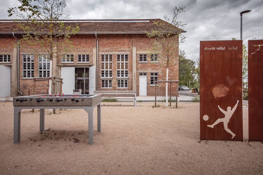 Das Foto zeigt den Außenbereich der Reithalle Achern, ein historisches Backsteingebäude mit großen Sprossenfenstern, das im Hintergrund zu sehen ist. Der Boden davor besteht aus hellem, feinem Kies. Im linken Vordergrund steht ein massiver Outdoor-Kickertisch aus grauem Stein oder Beton auf stabilen Metallfüßen. Rechts im Bild ragen zwei hohe, rost-braune Stahlstelen (Cortenstahl) auf. In die vordere Stele ist die Silhouette eines Fußballers und der Name „Julius Hirsch“ eingearbeitet. Hinter dem Kicker und zwischen jungen Bäumen lädt eine schlichte Bank zum Verweilen ein. Der Himmel ist bewölkt und taucht die Szenerie in ein weiches, gleichmäßiges Licht, das die industrielle Architektur betont.