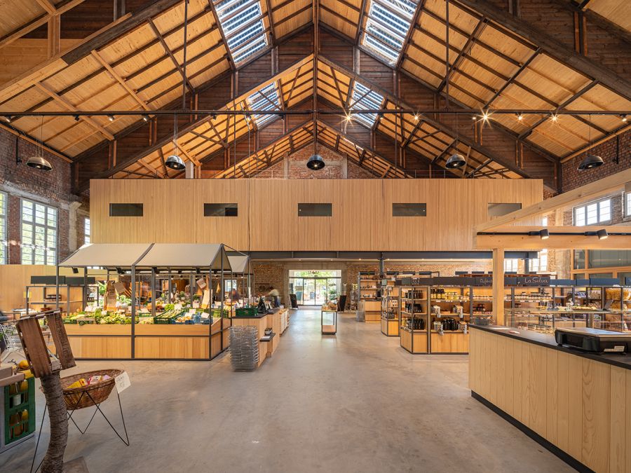 Das Foto zeigt die Markthalle Achern, die sich innerhalb der Reithalle befindet. Es ist eine weitläufige Halle mit einem markanten, offenen Holzdachstuhl. Das spitze Satteldach besitzt mehrere Dachfenster. Im Zentrum stehen moderne, helle hölzerne Verkaufsstände: Links werden in Körben und Kisten frisches Obst und Gemüse präsentiert. Rechts befinden sich Regale mit Delikatessen sowie eine Bedienungstheke. Im Hintergrund trennt eine obere Ebene aus hellem Holz den Raum ab. Der Boden besteht aus glattem, grauem Beton. Die Wände zeigen rotes Sichtbackstein-Mauerwerk. Die Atmosphäre wirkt durch das natürliche Licht und den warmen Holztönen einladend und modern.