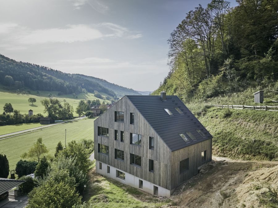Das moderne, mehrstöckiges Wohnhaus mit markanter Holzfassade steht an einem bewaldeten Hang. Das Gebäude hat eine asymmetrische Form: Eine Seite schließt mit einem steilen Pultdach ab, während die andere Fassade wie ein klassisches Giebelhaus wirkt. Die dunkle Holzverkleidung wird von zahlreichen quadratischen Fenstern unterschiedlicher Größe unterbrochen. Das Haus ist in eine idyllische Hügellandschaft eingebettet. Im Hintergrund erstreckt sich ein weites, grünes Tal mit Wiesen und vereinzelten Bäumen unter einem hellen, leicht dunstigen Himmel. Unmittelbar rechts hinter dem Gebäude steigt ein steiler, dicht bewaldeter Hang auf. Der Kontrast zwischen der geradlinigen, modernen Architektur und der sanften Natur prägt die Szenerie.