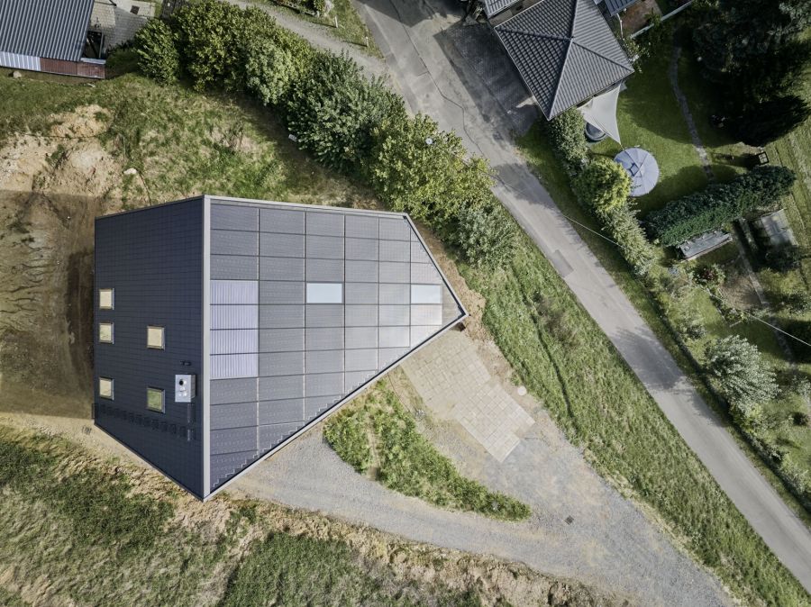 Die Luftaufnahme zeigt das moderne Gebäude mit einem außergewöhnlichen, asymmetrischen Dach in der Draufsicht. Die Dachfläche ist fast vollständig mit dunkelgrauen Photovoltaik-Modulen bedeckt, die in einem regelmäßigen Gitter angeordnet sind. Nur wenige kleine Dachfenster unterbrechen die Solarfläche. Das Haus hat eine unregelmäßige, fünfseitige Grundform und steht auf einem Grundstück mit hellbrauner Erde und grünen Grasflächen. Eine schmale Asphaltstraße verläuft diagonal am rechten Bildrand. In der Umgebung sieht man die Dächer benachbarter Häuser und dichte, grüne Bäume. Die Perspektive von oben betont die geometrische Form des Hauses und die effiziente Nutzung der Dachfläche für Solarenergie.