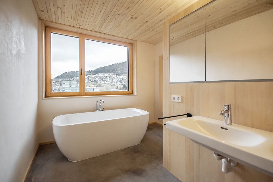 Das helle, minimalistisches Badezimmer ist überwiegend mit hellem Holz gestaltet. Im Zentrum steht eine freistehende, weiße Badewanne unter einem großen Holzfenster. Der Blick nach draußen zeigt eine hügelige Landschaft. Rechts im Vordergrund befindet sich ein Waschtisch aus hellem Holz mit einem schlichten weißen Waschbecken und einer modernen Armatur. Darüber hängt ein großer Spiegelschrank. Die Decke besteht aus hellen Holzpaneelen, während der Boden in einem kontrastierenden, dunklen Grau gehalten ist. Die Wände sind schlicht weiß verputzt. Die Atmosphäre wirkt durch das natürliche Licht und die warmen Materialien sehr einladend und ruhig.