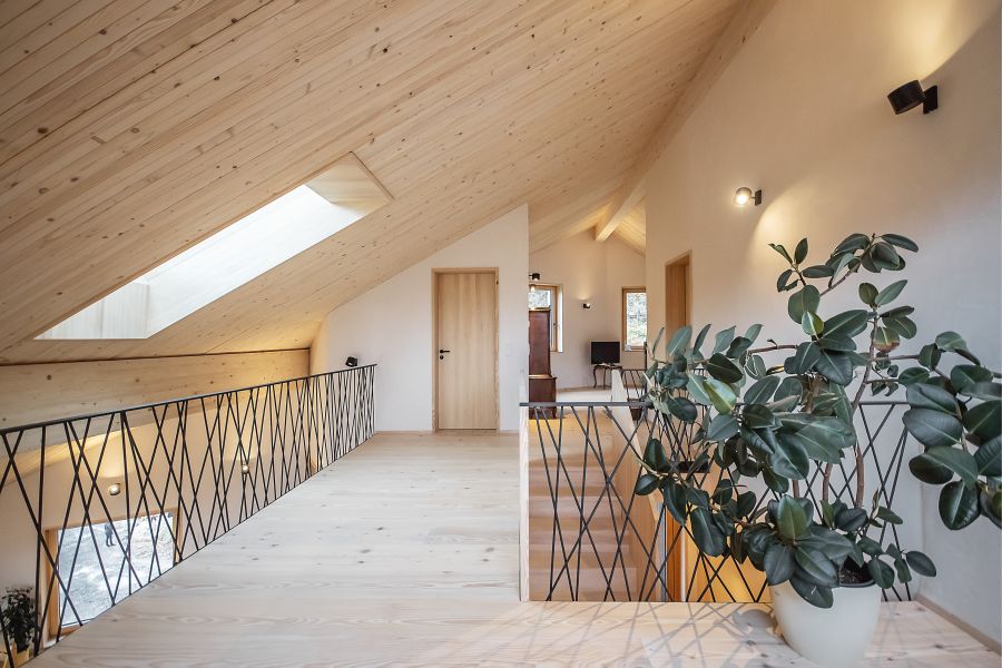 Ein heller, moderner Flur im Obergeschoss ist durch helles Holz und viel Licht geprägt. Die schräge Decke und der Boden bestehen aus hellem Massivholz. Ein langes Dachfenster flutet den Raum mit Tageslicht. Links begrenzt ein filigranes Geländer aus sich kreuzenden, schwarzen Metallstäben die Empore. Rechts führt eine Treppe nach unten, ebenfalls gesichert durch dieses dunkle Gitter. Im Vordergrund rechts steht eine große Zimmerpflanze mit dunklen, glänzenden Blättern in einem weißen Topf. An der weißen Wand gegenüber sind schlichte, schwarze Strahler angebracht. Im Hintergrund führt eine Holztür in einen weiteren Raum. Die Atmosphäre wirkt offen, warm und minimalistisch.