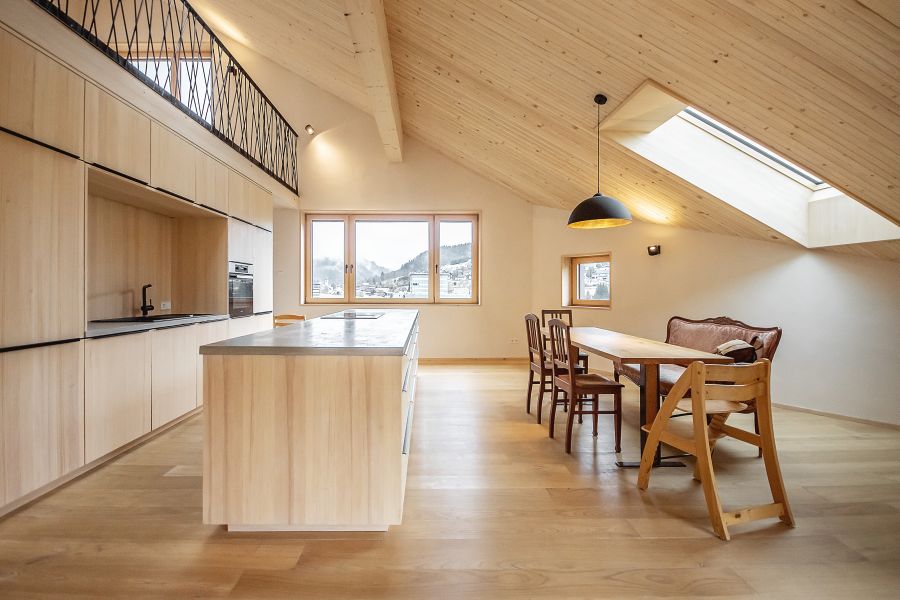 Ein lichtdurchfluteter, offen gestalteter Wohn- und Essbereich unter einer hohen Dachschräge aus hellem Holz. Links dominiert eine minimalistische Einbauküche mit hellen Holzfronten und einer großen Kochinsel. Ein filigranes, schwarzes Metallgeländer begrenzt eine Empore darüber. Rechts steht ein rustikaler Esstisch aus Holz mit einer Bank und klassischen Stühlen, sowie am vorderen Tischende ein Kinderstuhl aus hellem Holz. Ein großes Dachfenster und ein breites Frontfenster geben den Blick auf eine hügelige Landschaft frei. Eine schwarze Hängeleuchte setzt einen modernen Akzent über dem Tisch. Der durchgehende Holzboden und die weißen Wände verstärken die warme, natürliche Atmosphäre des Raumes.