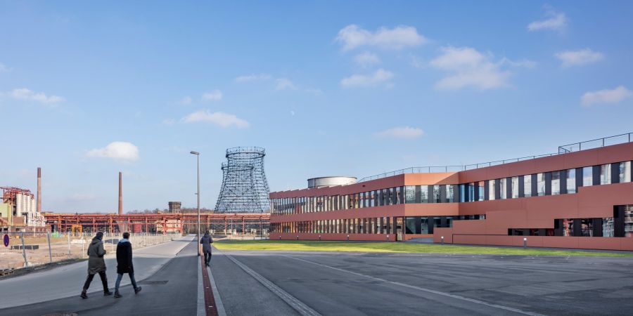 Weitläufiges Industriegelände unter hellblauem, leicht bewölktem Himmel. Im Fokus stehen der Kontrast zwischen moderner Architektur und historischer Industrie. Rechts: Ein langgestrecktes, zweistöckiges Bürogebäude mit rötlich-brauner Fassade (terrakottafarben) und horizontalen Fensterbändern, die das Tageslicht spiegeln. Mitte/Hintergrund: Die filigrane, offene Stahlgitter-Konstruktion eines alten Kühlturms ragt empor. Links: Eine Asphaltfläche mit einem Fußweg, auf dem drei Personen vom Betrachter weggehen (zwei nebeneinander, eine weiter vorne). Im fernen Hintergrund stehen flache Industriegebäude und zwei dünne Schornsteine. Die Szene ist hell erleuchtet, wirkt aufgeräumt und ruhig.