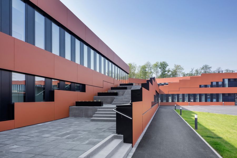 Das Bild zeigt den modernen Außenbereich eines mehrstöckigen Gebäudekomplexes mit einer markanten, terrassenförmigen Architektur. Fassade: Die Wände sind mit großen, matt-terrakottafarbenen Platten verkleidet. Lange, horizontale Fensterbänder mit schwarzen Rahmen unterbrechen die farbigen Flächen. Wege und Treppen: Im linken Vordergrund führt eine breite Betontreppe nach oben. Sie wird von schwarzen Geländern und terrakottafarbenen Brüstungen flankiert, die stufenförmig ansteigen. Der Boden davor ist mit grauen quadratischen Platten gepflastert. Rechter Bereich: Ein asphaltierter Fußweg verläuft parallel zum Gebäude. Daneben befindet sich eine gepflegte Rasenfläche mit kleinen, modernen Pollerleuchten. Im Hintergrund öffnet sich ein asphaltierter Hof.