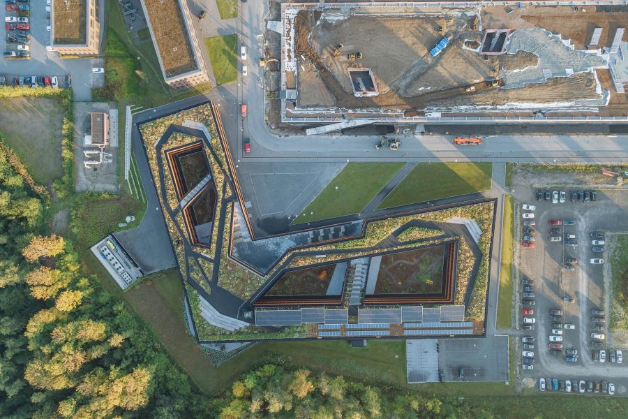 Eine senkrechte Luftaufnahme zeigt das moderne Bürogebäude der RAG-Stiftung auf Zollverein aus der Vogelperspektive. Das langgestreckte Bauwerk in L-Form besticht durch seine markante, geometrische Architektur mit mehreren Winkeln und integrierten Innenhöfen. Das Flachdach ist vollständig begrünt und mit Rasenflächen, Wegen sowie Reihen von bläulich schimmernden Photovoltaikanlagen gestaltet. Links und unten grenzt der Gebäudekomplex an dichte, grüne Baumkronen eines Waldes. Am oberen Bildrand verläuft eine Straße, hinter der eine größere Baustelle mit einigen Baufahrzeugen liegt. Rechts befindet sich ein asphaltierter Parkplatz mit zahlreichen Autos. Das warme Sonnenlicht wirft lange, weiche Schatten nach links oben.