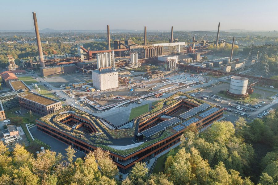 Die Luftaufnahme zeigt ein modernes, verwinkeltes Bürogebäude mit intensiv begrüntem Dach im Vordergrund. Das Gebäude umschließt mehrere Innenhöfe und ist mit Solaranlagen ausgestattet. Es grenzt an ein dichtes, herbstlich gefärbtes Waldstück. Im Hintergrund erstreckt sich eine riesige, historische Industrieanlage mit einer Kokerei. Zu sehen sind markante, hohe Industrieschornsteine, rostbraune Rohrleitungssysteme, Kühltürme und große Fabrikhallen aus Backstein und Stahl. Zwischen dem modernen Neubau und der alten Industriekulisse befinden sich Freiflächen, Baustellenbereiche und Parkplätze. Die Szenerie ist in warmes, helles Tageslicht getaucht, was einen starken Kontrast zwischen der grünen Natur, der modernen Architektur und dem industriellen Erbe schafft.