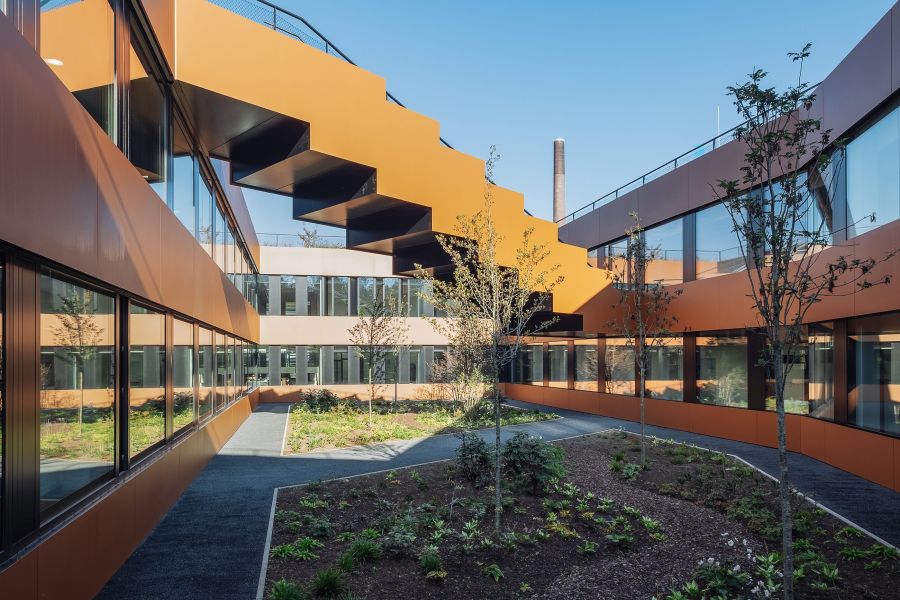 Die Fotografie zeigt einen modernen Innenhof des Bürogebäudes, geprägt von geometrischer Architektur und warmen Farbtönen. Das mehrstöckige Gebäude umschließt den Hof mit durchgehenden Fensterfronten, die den Blick auf gegenüberliegende Büros freigeben. Die Fassadenelemente erstrahlen in einem matten, rötlich-braunen Kupfer- oder Cortenstahl-Ton. Ein markantes Gestaltungselement ist eine skulpturale ansteigende Außentreppe mit geschlossener, kupferfarbener Brüstung. Sie spannt sich diagonal über einen Teil des Hofes und verbindet die oberen Stockwerke. Der Boden des Innenhofs ist mit Beeten, Rindenmulch und jungen, schlanken Bäumen bepflanzt. Ein grauer, asphaltierter Fußweg schlängelt sich durch das Grün. Im Hintergrund ragt ein einzelner Industrieschornstein in den wolkenlosen, blauen Himmel.