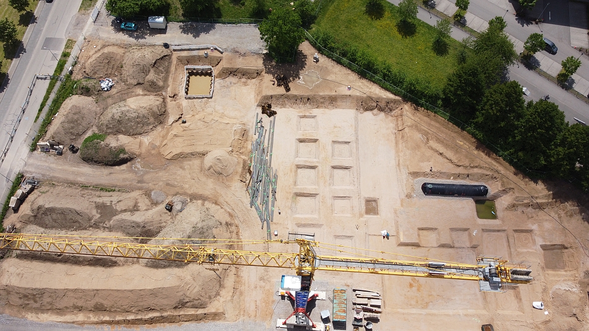 Drohnenflug im Mai über die Baustelle. Im Vordergrund ist der Kran von oben zu sehen, rechts der Aushub für den Laborbau. Dahinter befindet sich ein schwarzer Regenwassertank.