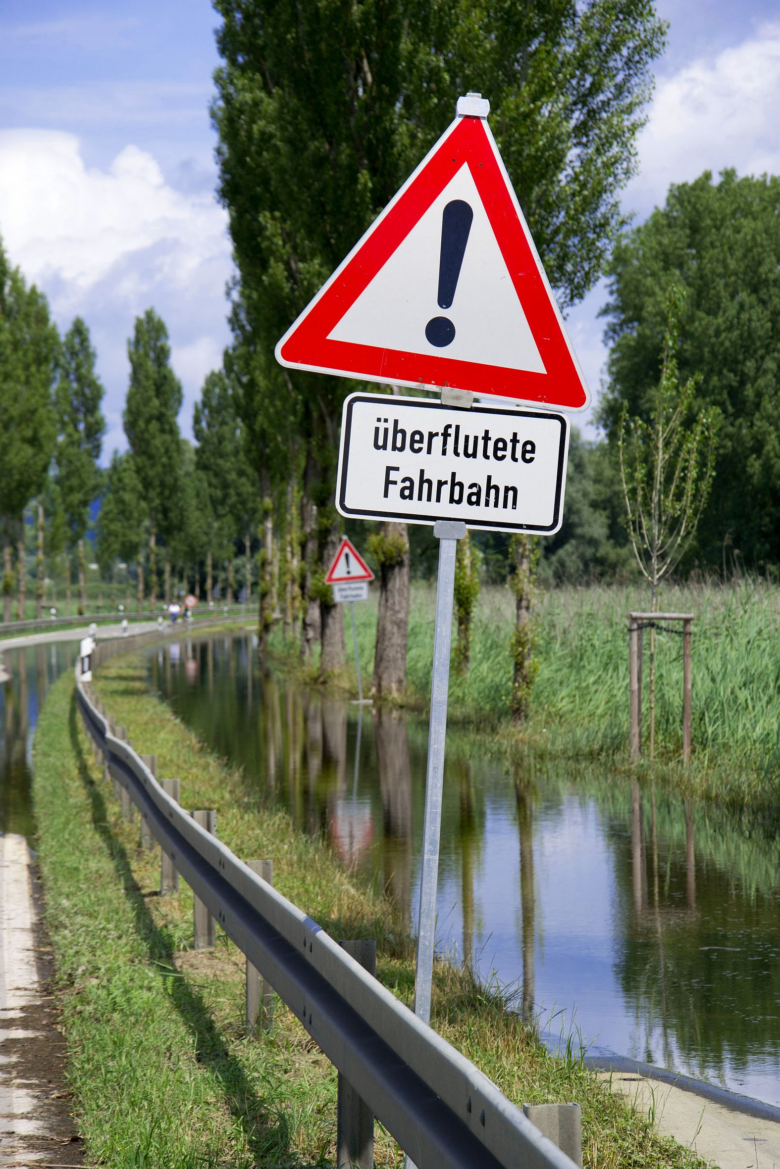 Ein Schild mit der Aufschrift "Überflutete Fahrbahn", im Hintergrund eine Allee mit überluteter Straße