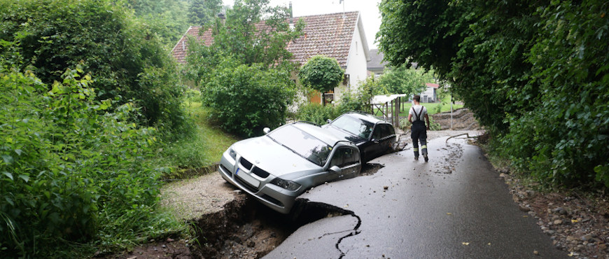  Das Bild zeigt eine durch starke Regenfälle im Juni 2021 unterspülte und dadurch aufgerissene Straße in der Gemeinde Küssaberg in Baden-Württemberg. Zwei Autos, die dort geparkt wurden, sind durch den entstandenen Riss in den Grund gerutscht.